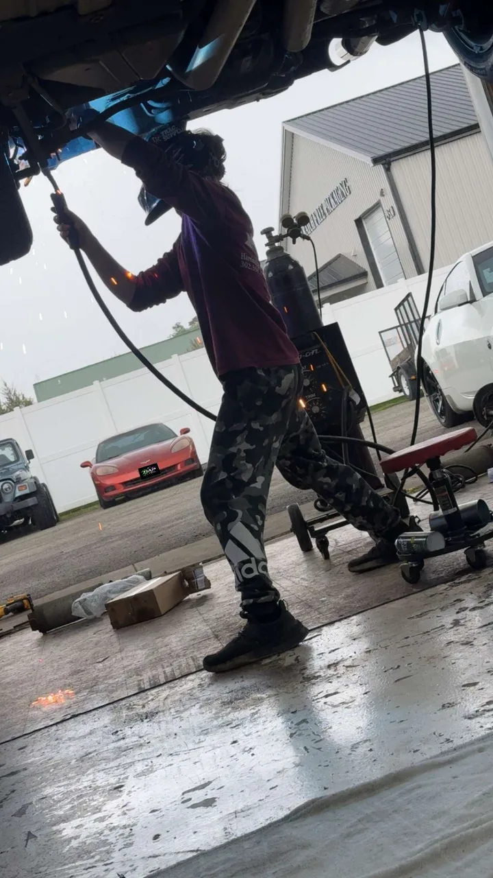 A person working beneath a raised vehicle in the shop; background license plate covered